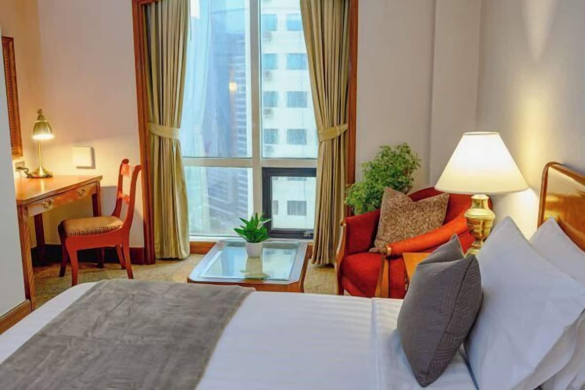 A hotel room with a bed, a desk, a chair, a small table with a plant, and a view of buildings through the window.