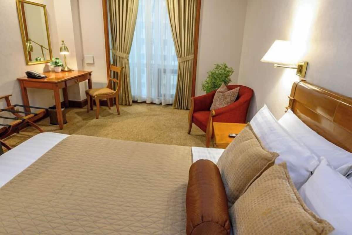 A hotel room with a large bed, a desk with a chair, a red armchair, and a lamp.