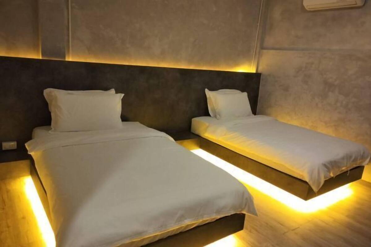 Two beds with white linens and underfoot lighting, in a room with a textured wall and a dark headboard.
