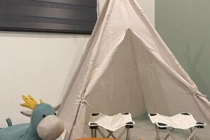 Une structure en forme de tente, recouverte d’un tissu doux et de couleur claire, un jouet en peluche représentant un cheval bleu avec une crinière jaune, et deux petites chaises pliantes avec des assises en tissu blanc.