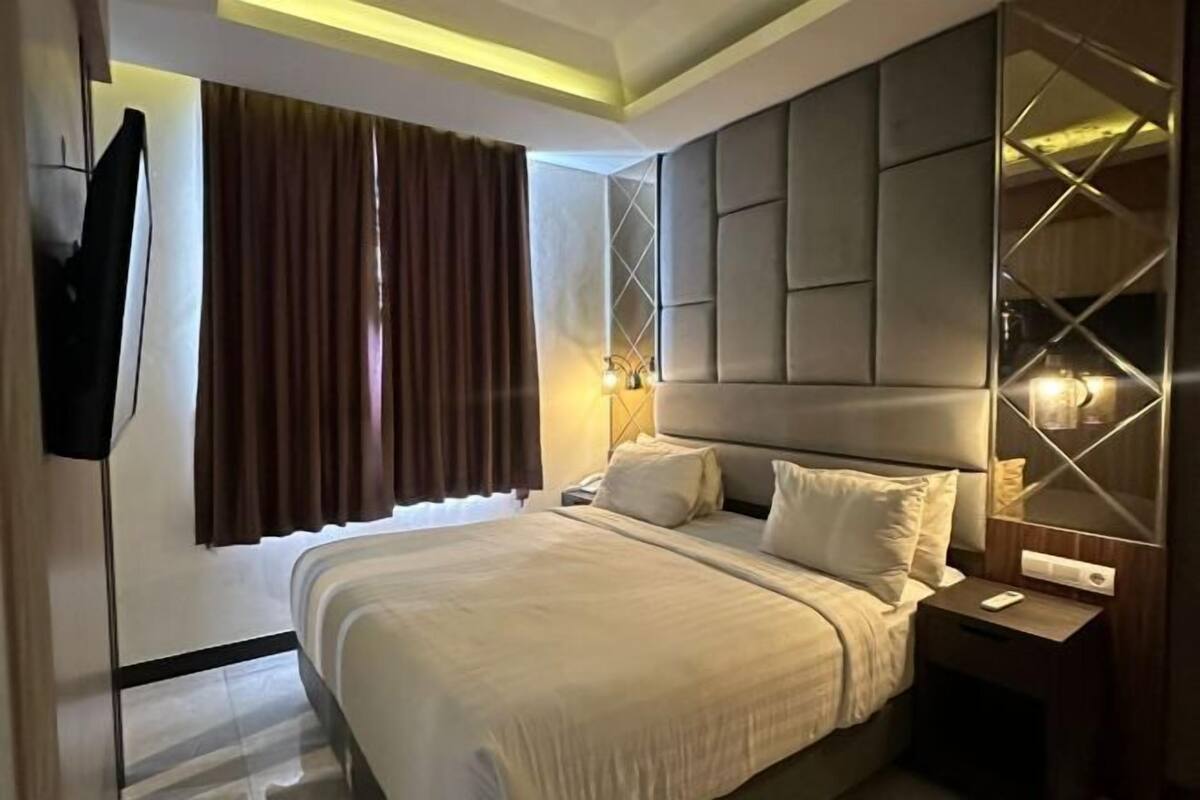 Une chambre d’hôtel moderne avec un grand lit, une table de chevet, un téléviseur fixé au mur et une grande fenêtre avec des rideaux.