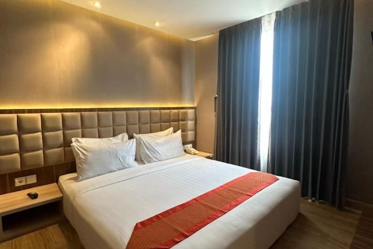A hotel room with a large bed, a headboard, a bedside table, and a window with curtains.
