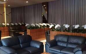 A modern lounge area with leather sofas, a wooden planter with white flowers, and a staircase in the background.