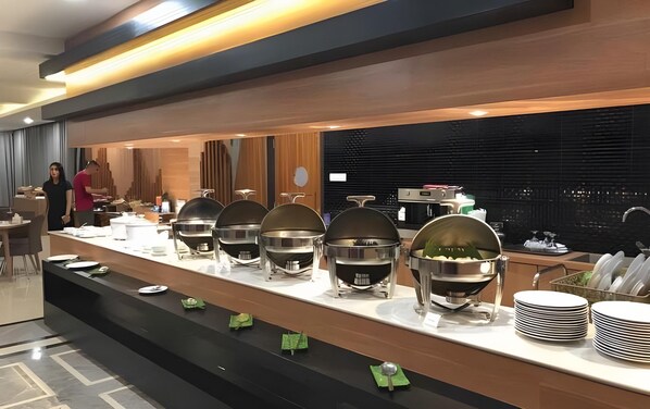 A buffet setup with multiple serving stations, a counter with stacked plates, and a view of the city through large windows.