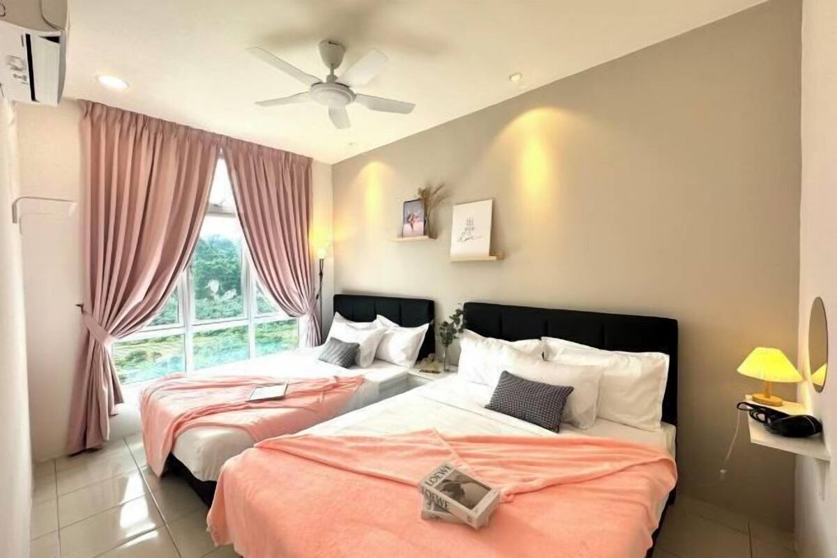 A hotel room with two beds, a ceiling fan, a window with curtains, a bedside table with a lamp, and a wall-mounted shelf with a mirror.