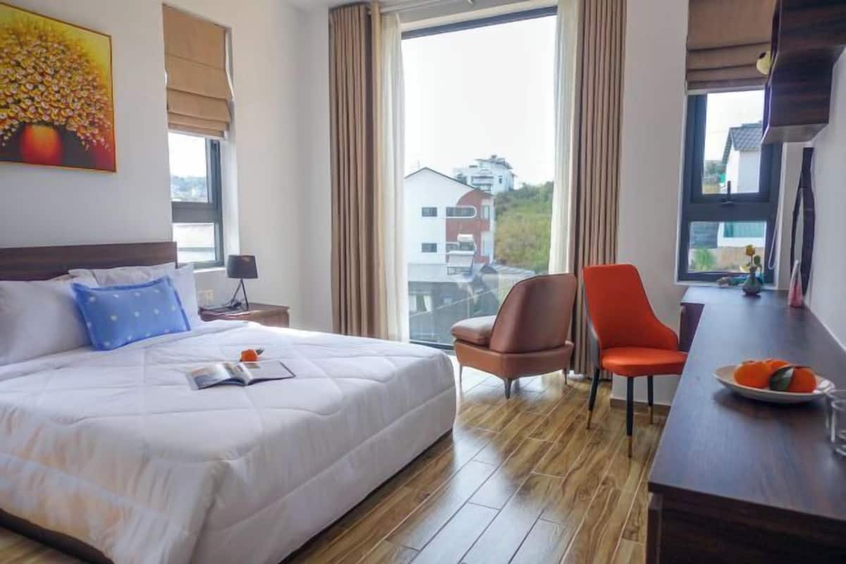 A hotel room with a large bed, two chairs, a desk, and a view of buildings outside.