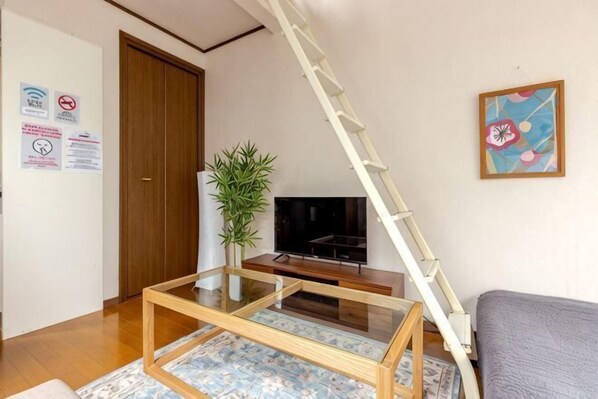 Een woonkamer met een televisie, een plant, een ladder en een schilderij aan de muur.