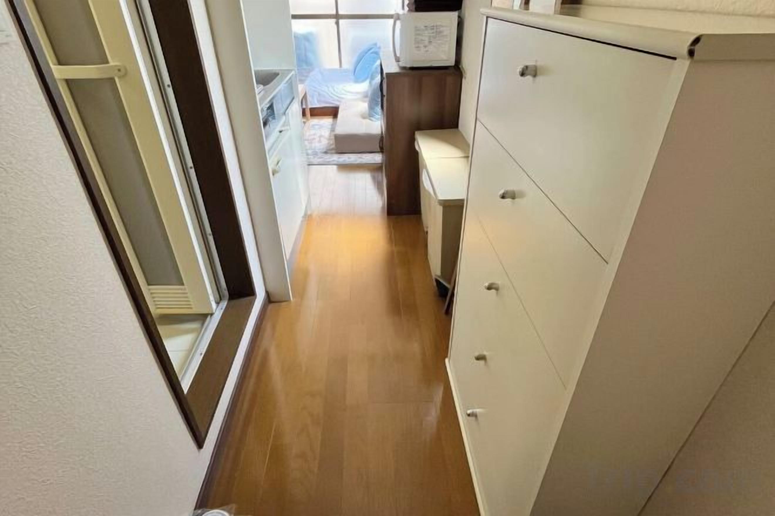 A narrow hallway with a wooden floor leading to a room with a bed and a window.