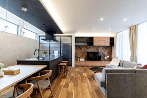 Uma cozinha moderna com uma ilha central, sala de jantar com cadeiras de madeira, sofá e televisão ao fundo.