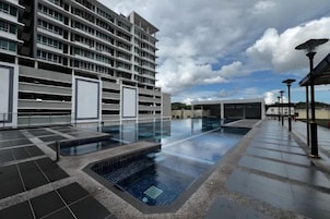 A hotel pool area with a modern building, a covered seating area, and a clear sky.