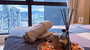 Habitación de hotel con un ventanal grande, una cama con una toalla enrollada, una botella de agua y un plato con flores secas.