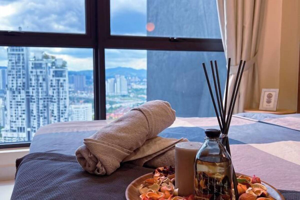 Une chambre d’hôtel avec une grande fenêtre, un lit sur lequel est posée une serviette roulée, une bouteille d’eau et une assiette de fleurs séchées.