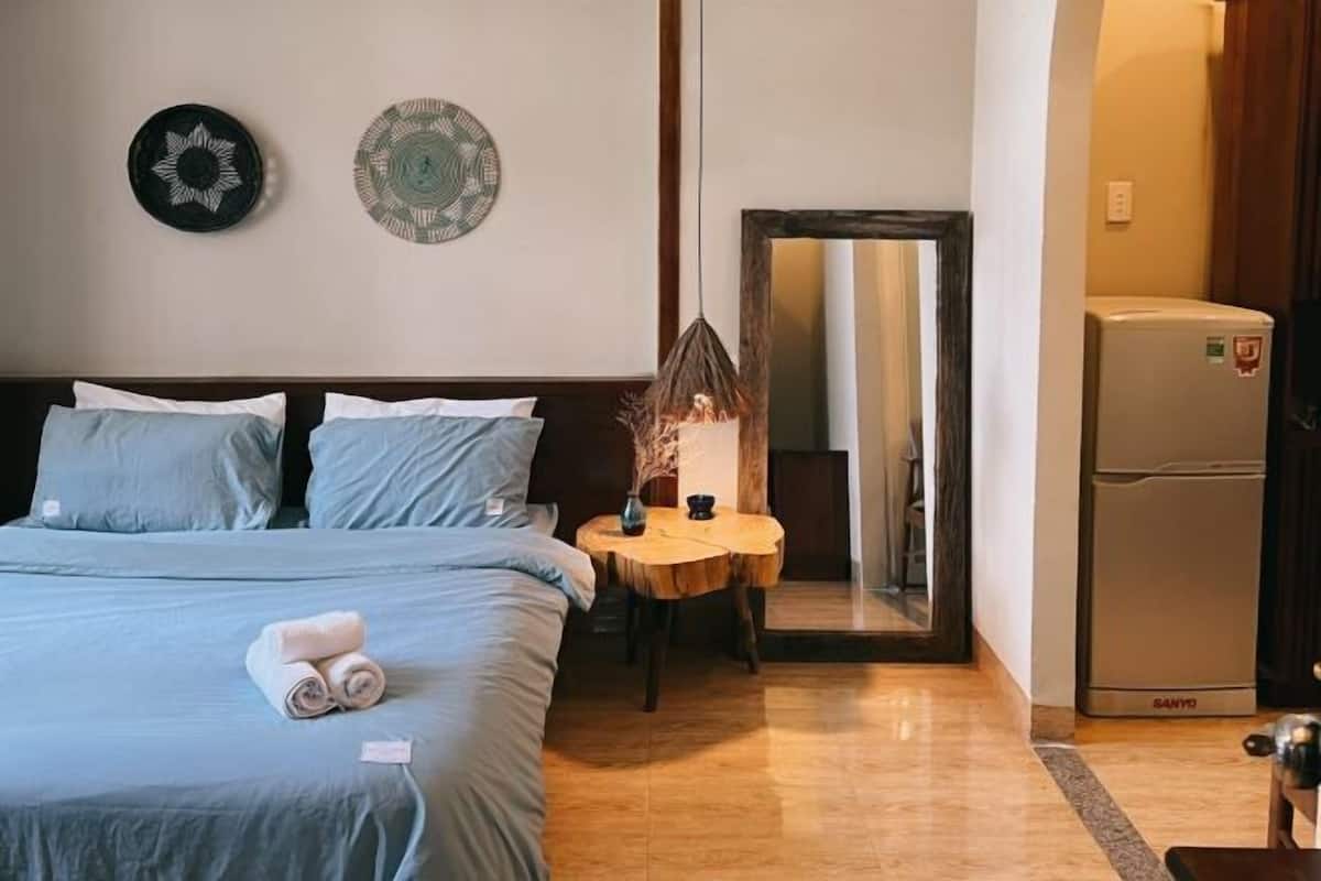 A hotel room with a bed, a small table with a vase, a mirror, and a refrigerator.