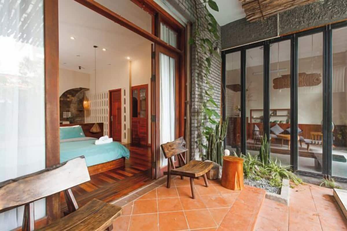 A room with a bed, a wooden chair, a small table, and a potted plant.