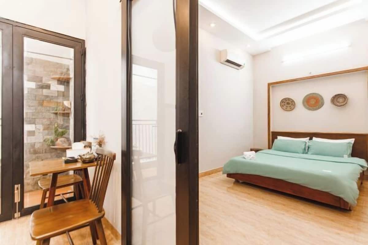 A bedroom with a bed, a wooden floor, a dining area with a table and chairs, and a view through a glass door.