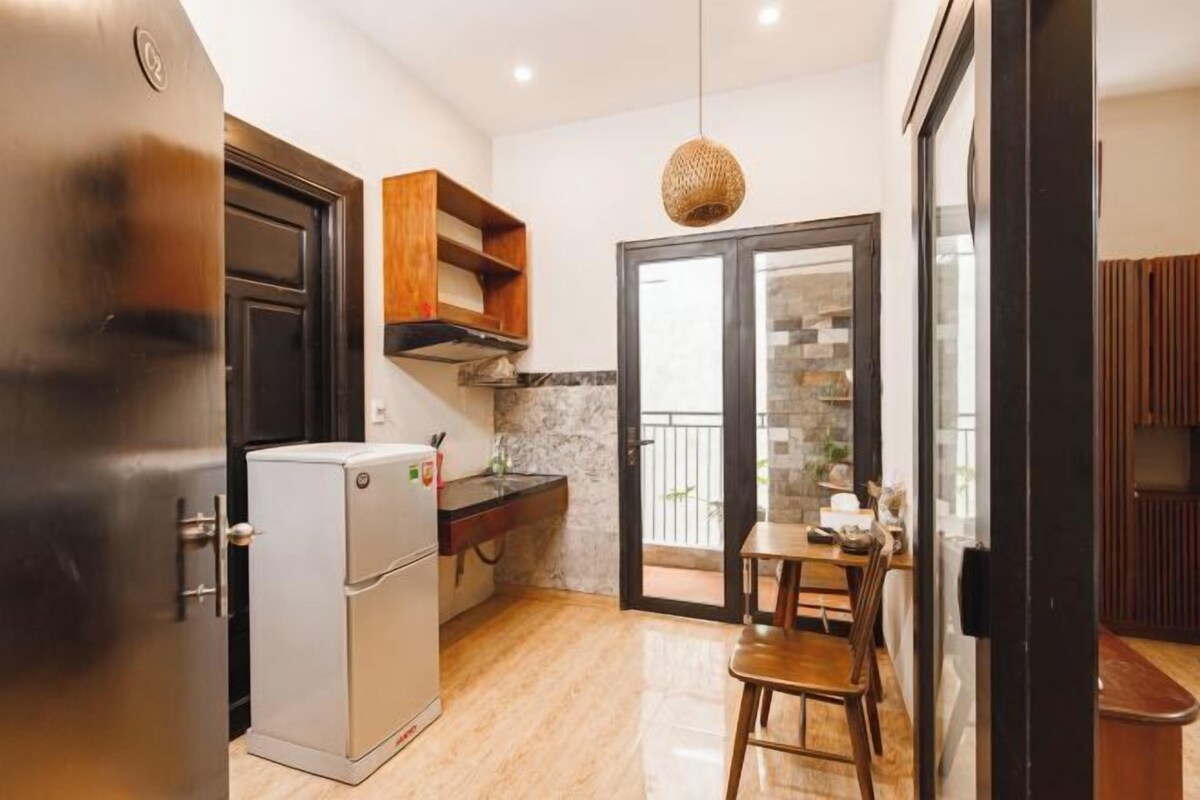 A compact kitchen with a refrigerator, wooden cabinets, a hanging light fixture, and a sliding glass door leading to a balcony.