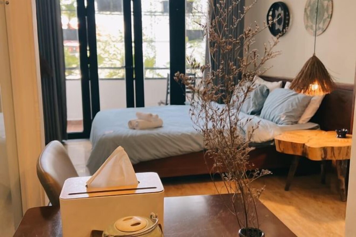 A hotel room with a bed, a bedside table with a lamp, a chair, and a window with curtains.