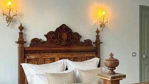 Un lit bien fait, avec du linge de lit blanc, une tête de lit en bois ornée de motifs complexes et deux appliques murales diffusant une lumière chaleureuse.