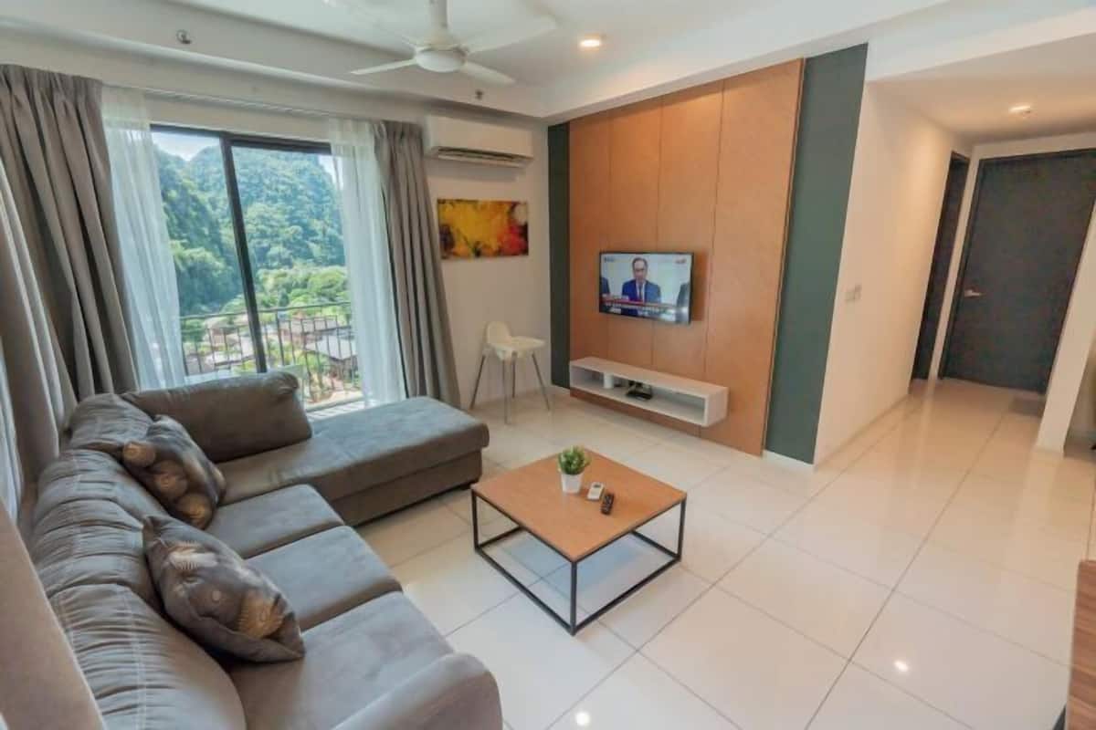 A modern living room with a sofa, a coffee table, a TV mounted on the wall, and a view of greenery outside.