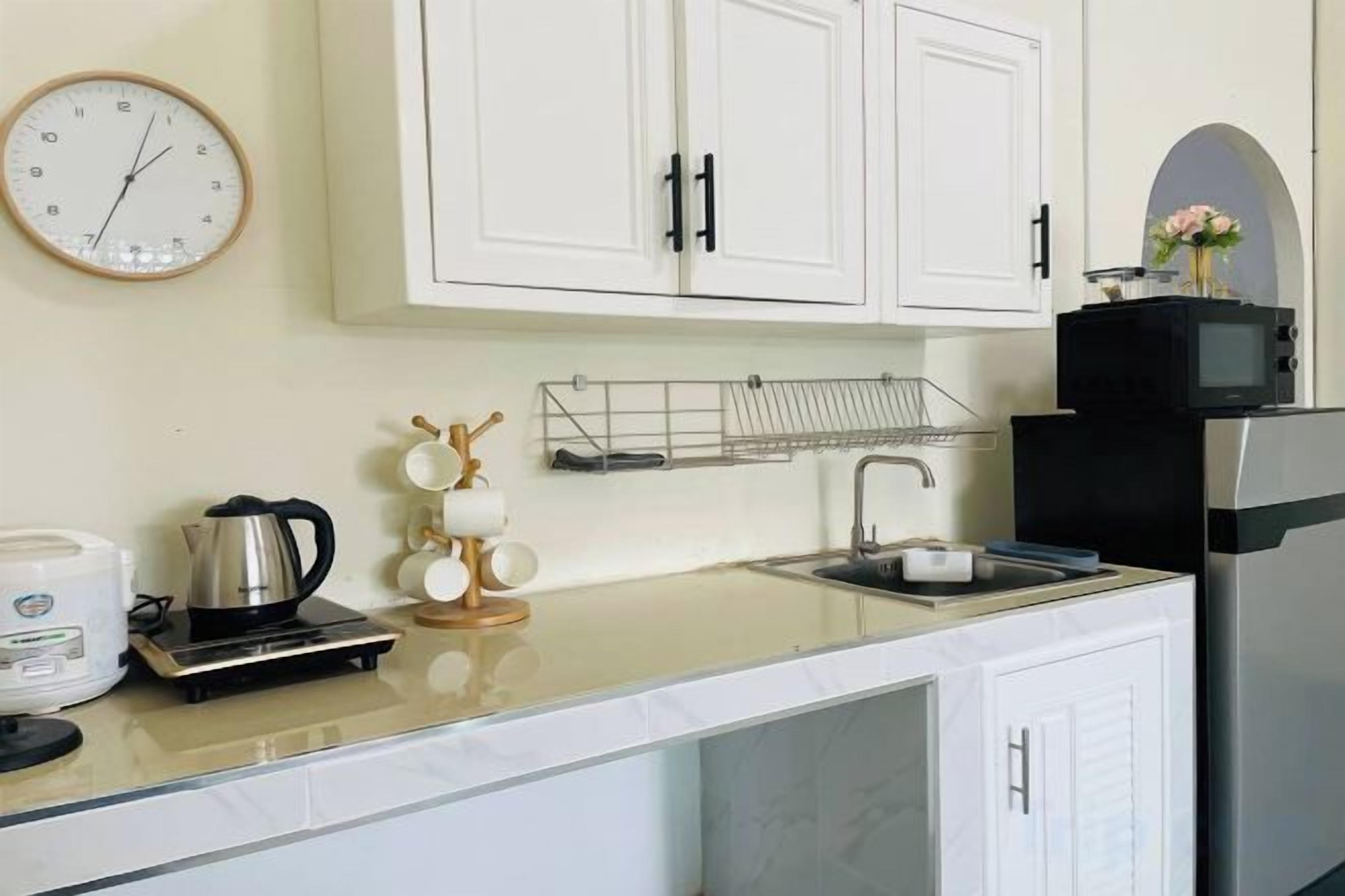 A kitchen with white cabinets, a microwave, a kettle, and a clock on the wall.