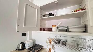 Una cocina con microondas, hervidor y otro hervidor sobre la encimera. Hay una estantería con platos y un escurreplatos con utensilios.