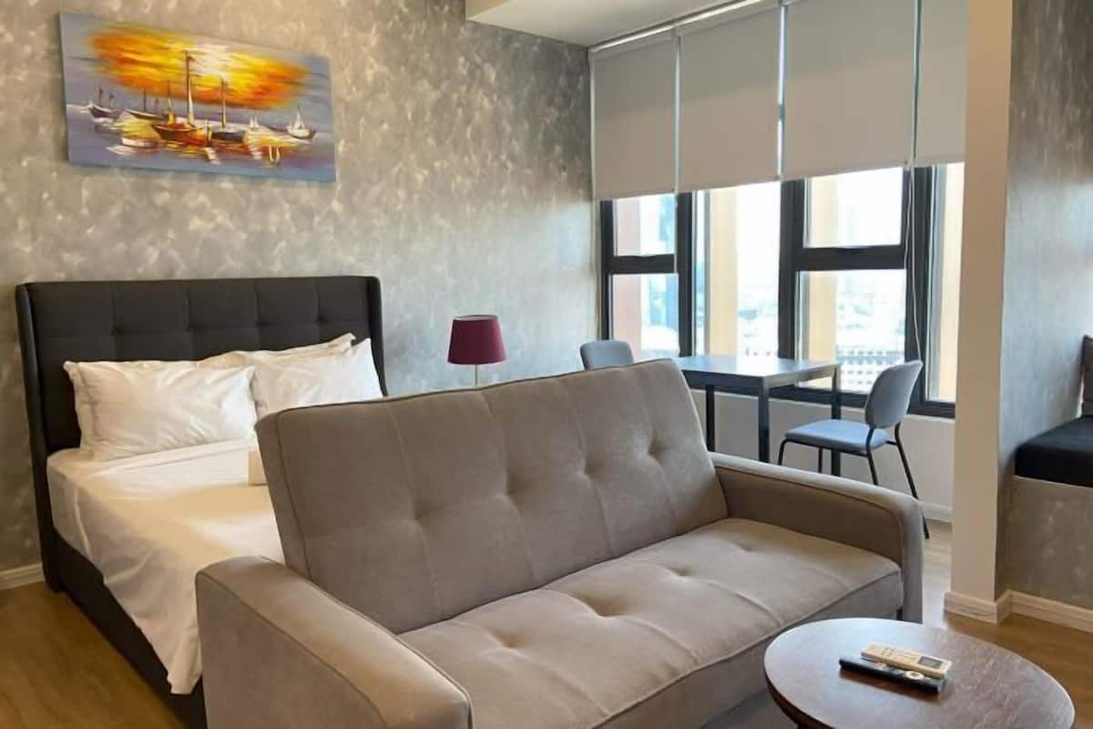 A modern hotel room with a bed, sofa, and a view of the city through large windows.
