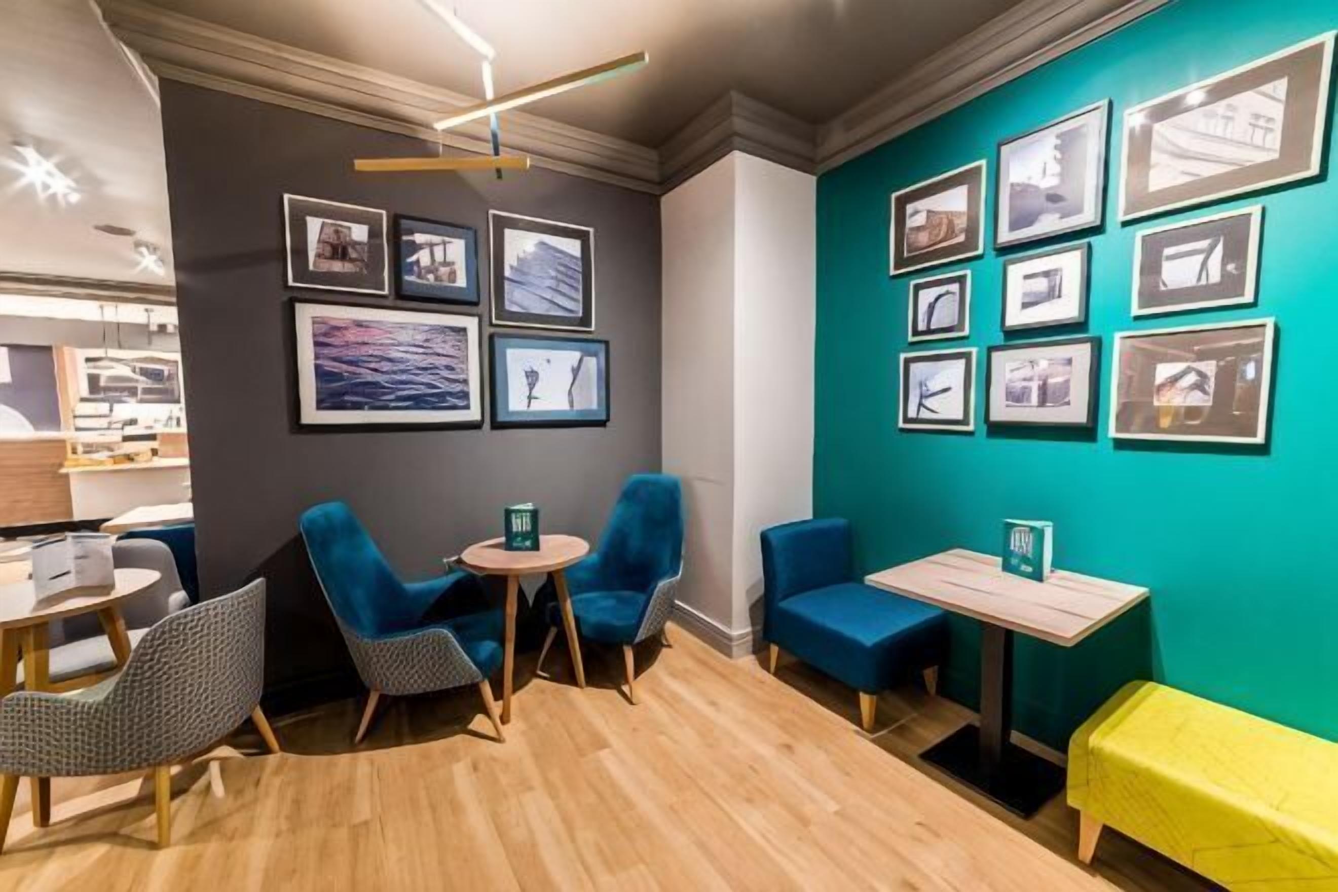 A modern hotel room with a wooden floor, blue armchairs, a small round table, and a yellow bench.
