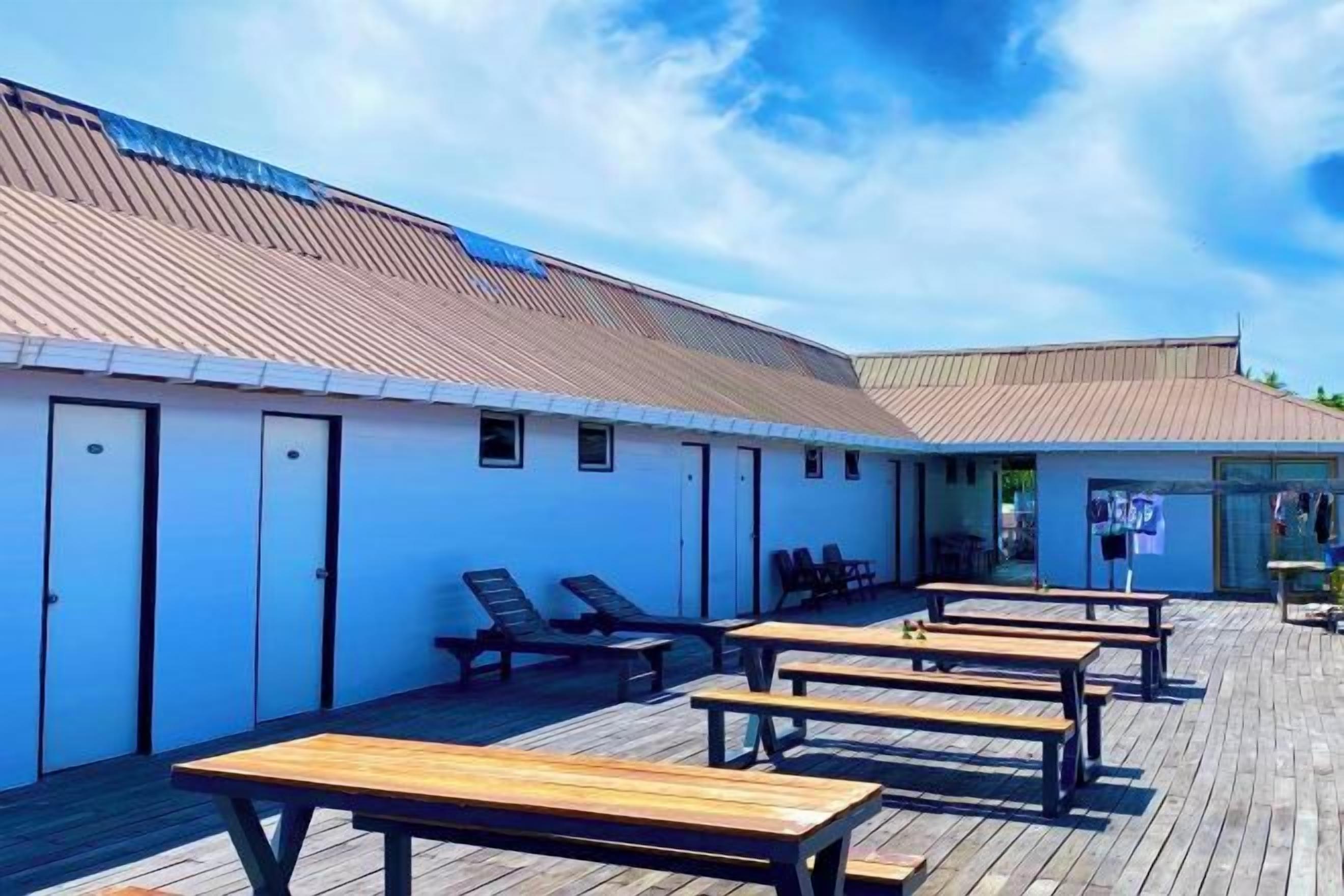 Un'area esterna coperta con panche e tavoli in legno, un edificio con tetto spiovente e cielo sereno.