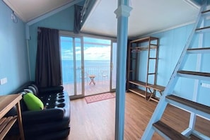 A room with a large sliding glass door leading to a balcony with a view of the sea. There is a black sofa with a green pillow and a wooden table.