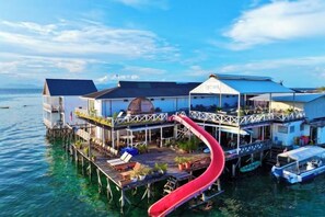 Una casa flotante con un tobogán rojo, estructura sobre el agua, y un cielo azul cristalino.