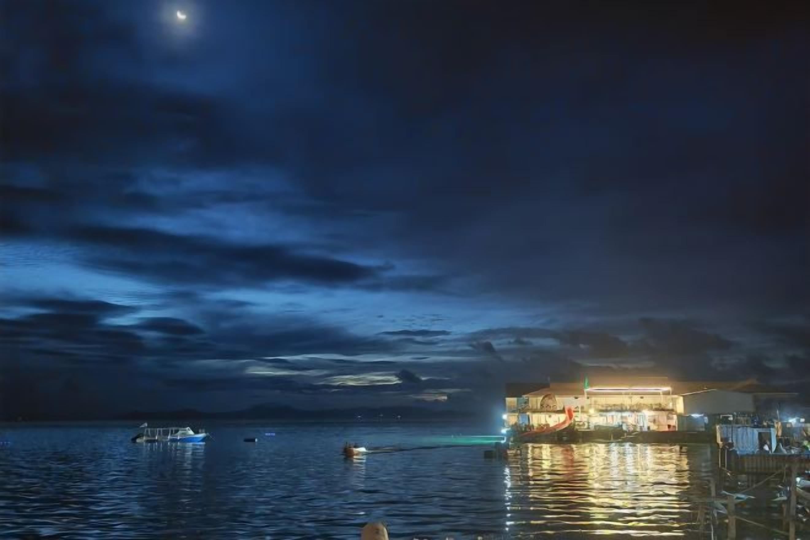 A waterfront hotel with a large illuminated building, calm water, and a boat in the distance.