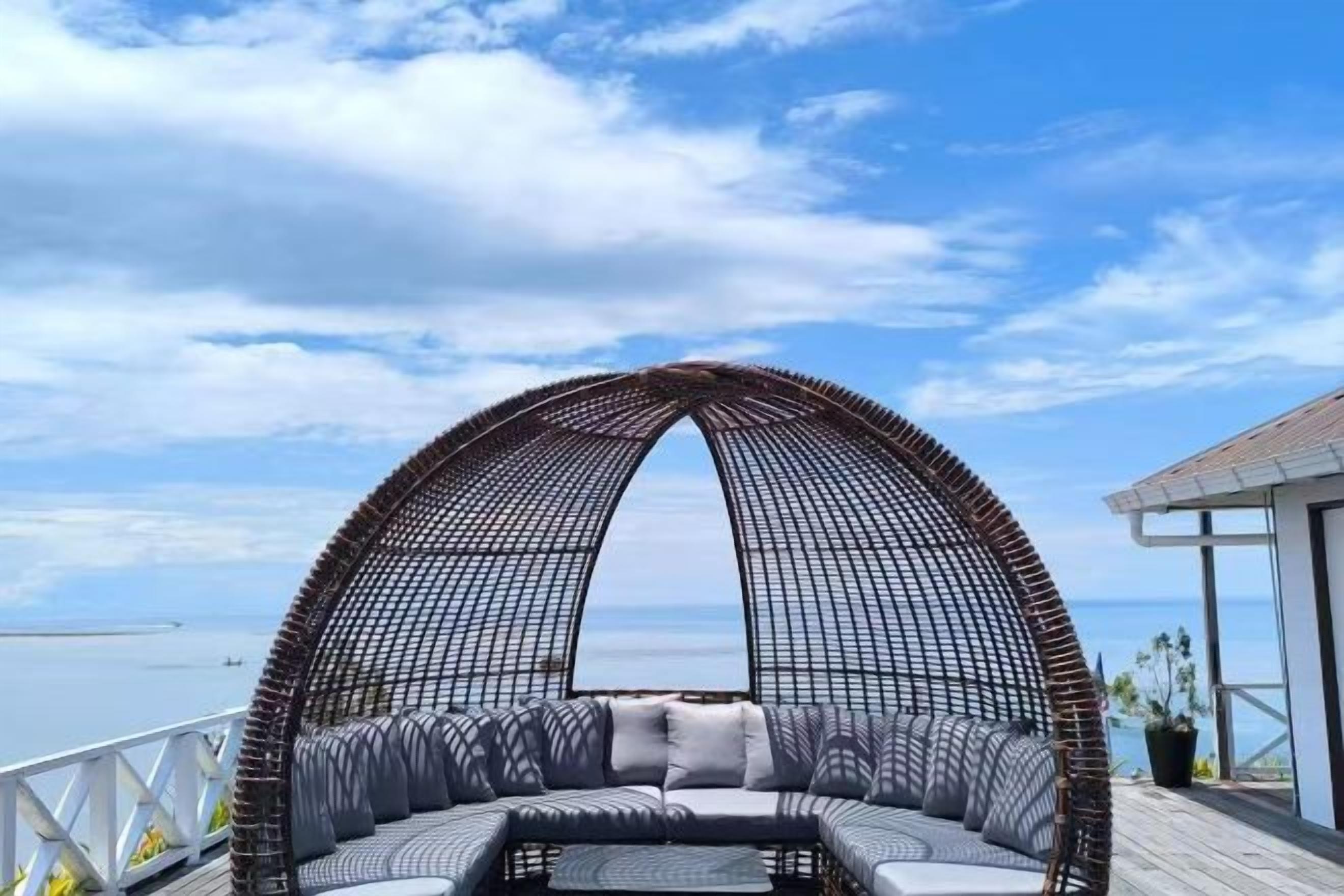 Een ronde rieten bank met kussens, geplaatst op een houten terras met uitzicht op zee.