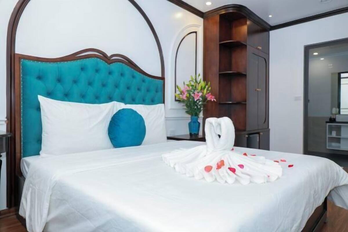 A hotel room with a bed, a blue headboard, a white bedspread, a white pillow, a white towel, and a vase with flowers.
