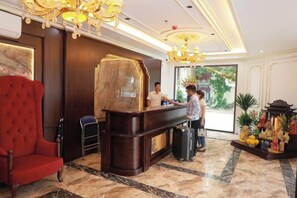 Een hotel receptie met een marmeren vloer, een rode fauteuil, een receptiebalie en een decoratief altaar met offers.