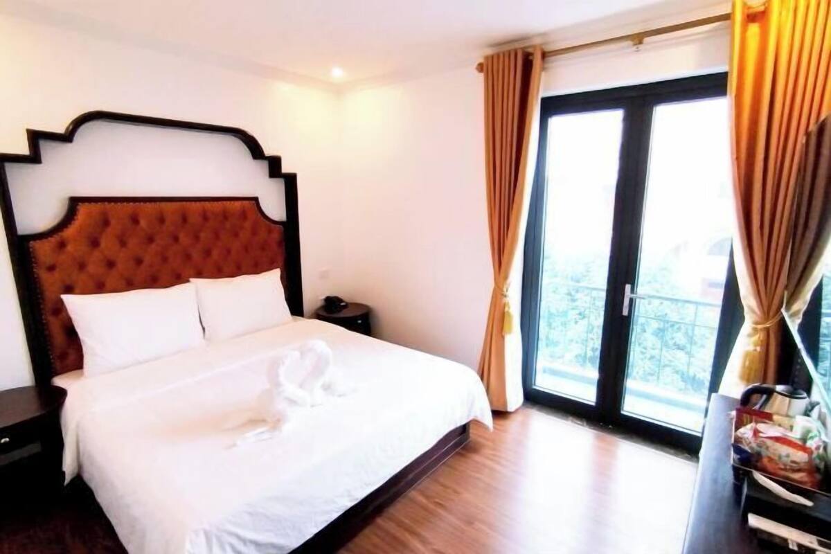 A hotel room with a large bed, a wooden headboard, a nightstand, and a view of the outdoors through a large window.