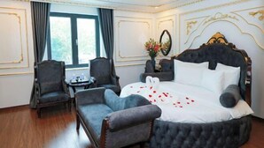Een slaapkamer met een groot bed, twee fauteuils, een klein tafeltje en een raam met gordijnen.