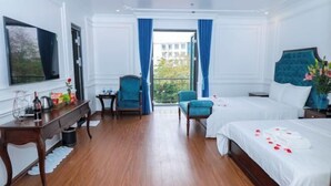 Hotelkamer met houten vloer, een bed met witte lakens, een bureau met stoel, een kleine tafel met stoel, en een groot raam met blauwe gordijnen.