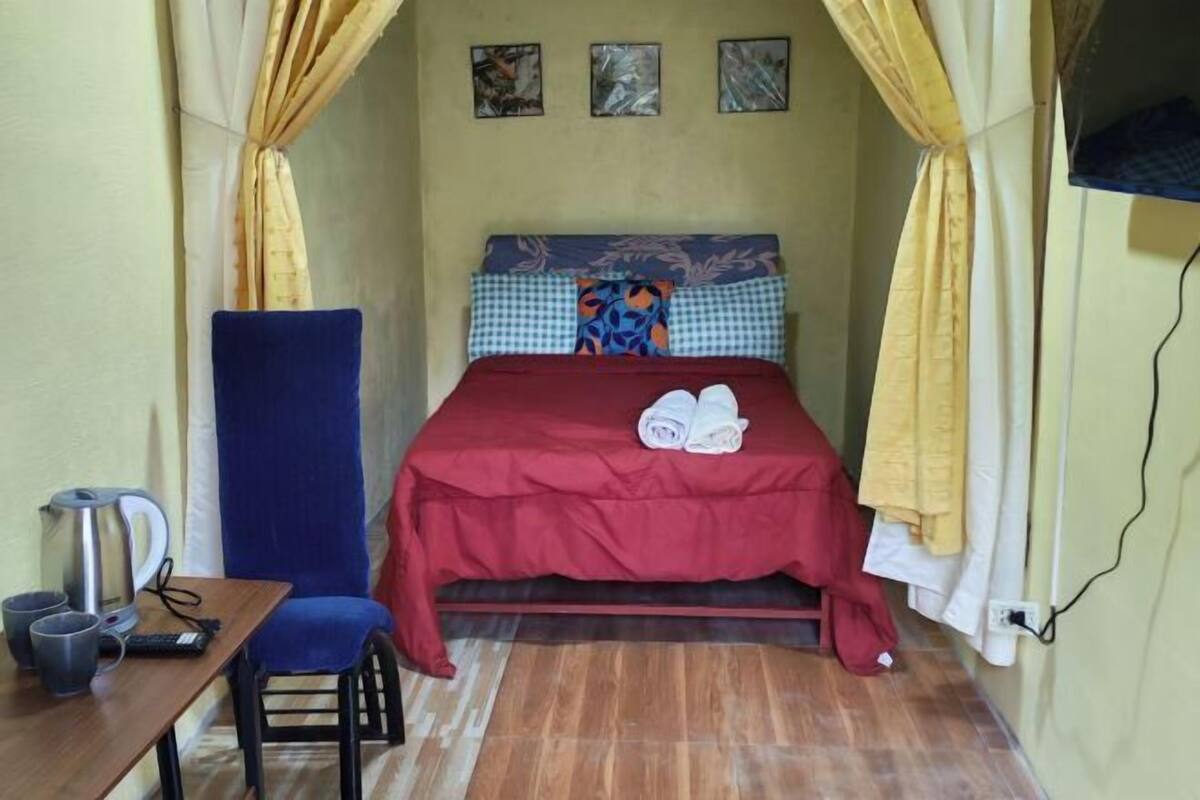 A small room with a bed, a desk with a kettle, a chair, and a blue curtain.
