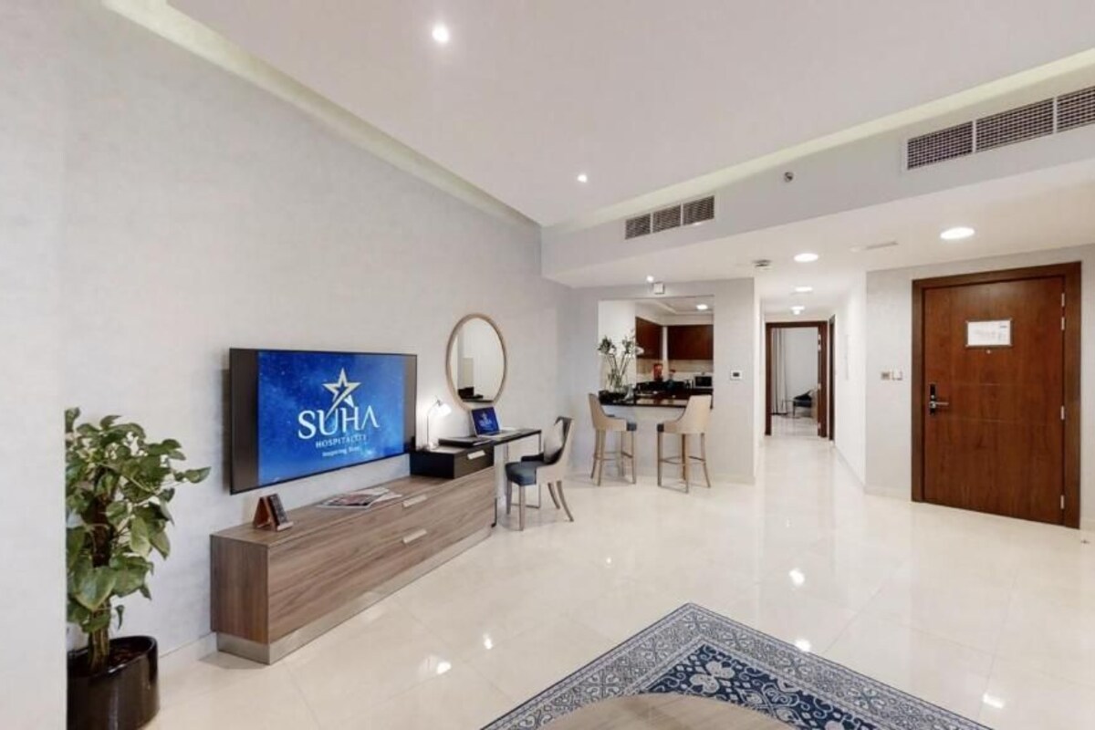 A modern hotel lobby with a flat-screen TV, a reception desk, a dining area, and a kitchen in the background.