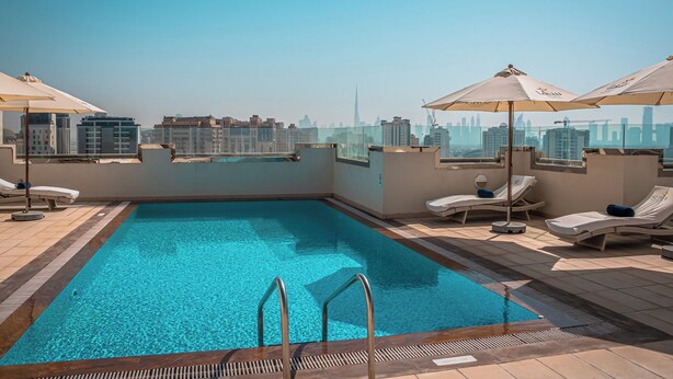 A rooftop pool with clear blue water, sun loungers, and umbrellas, overlooking a city skyline.