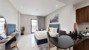 A modern hotel room with a bed, desk, chair, and kitchenette.