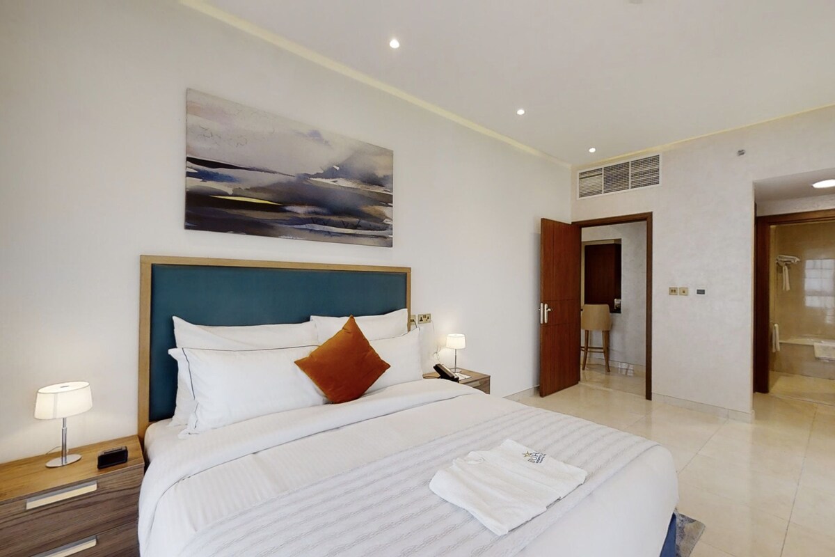 A hotel room with a large bed, a headboard, a bedside table with a lamp, a painting on the wall, and a bathroom door.