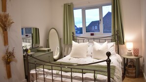Ein Schlafzimmer mit Bett, Kommode, Spiegel, Fenster mit Vorhängen und Nachttisch mit Lampe.