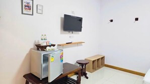 A compact living space with a refrigerator, a small wooden table, a TV mounted on the wall, and a shelf with a few items. - Khunnampheung Apartment (Sakon Nakhon)
