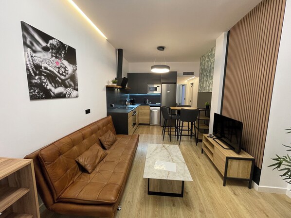 Un salon moderne avec un canapé en cuir marron, une table basse et un téléviseur posé sur un meuble en bois. On aperçoit au fond une cuisine équipée d’un four à micro-ondes, d’un réfrigérateur et d’un évier.