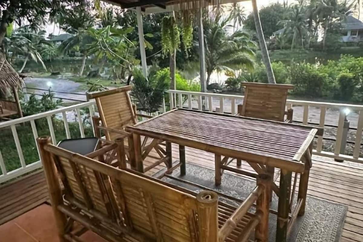 Terraza de madera con mesa y sillas, rodeada por una barandilla blanca, con vista a un jardín tropical y un cuerpo de agua.