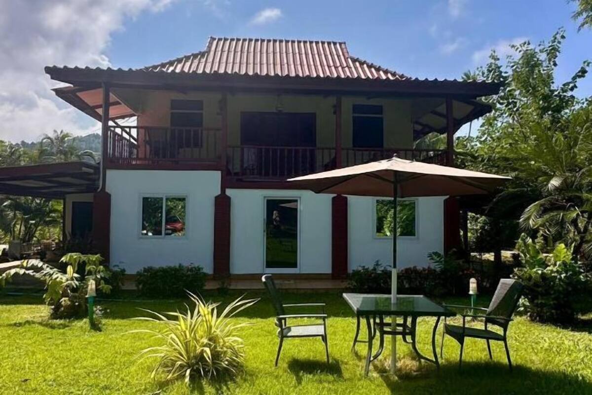 Una casa de dos pisos con un balcón, un patio con mesa y sillas, y un jardín.