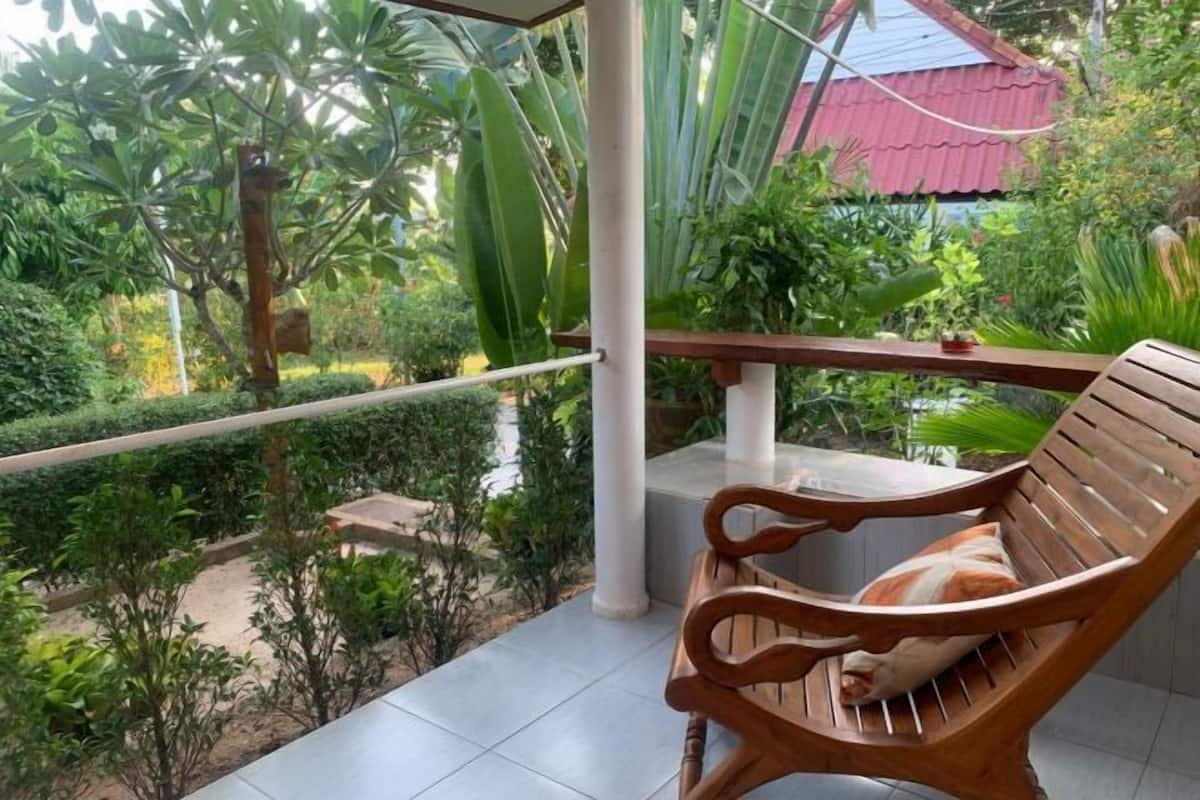 Un banco de madera con cojín en un balcón con baldosas, con vistas a un jardín y un edificio de tejado rojo.