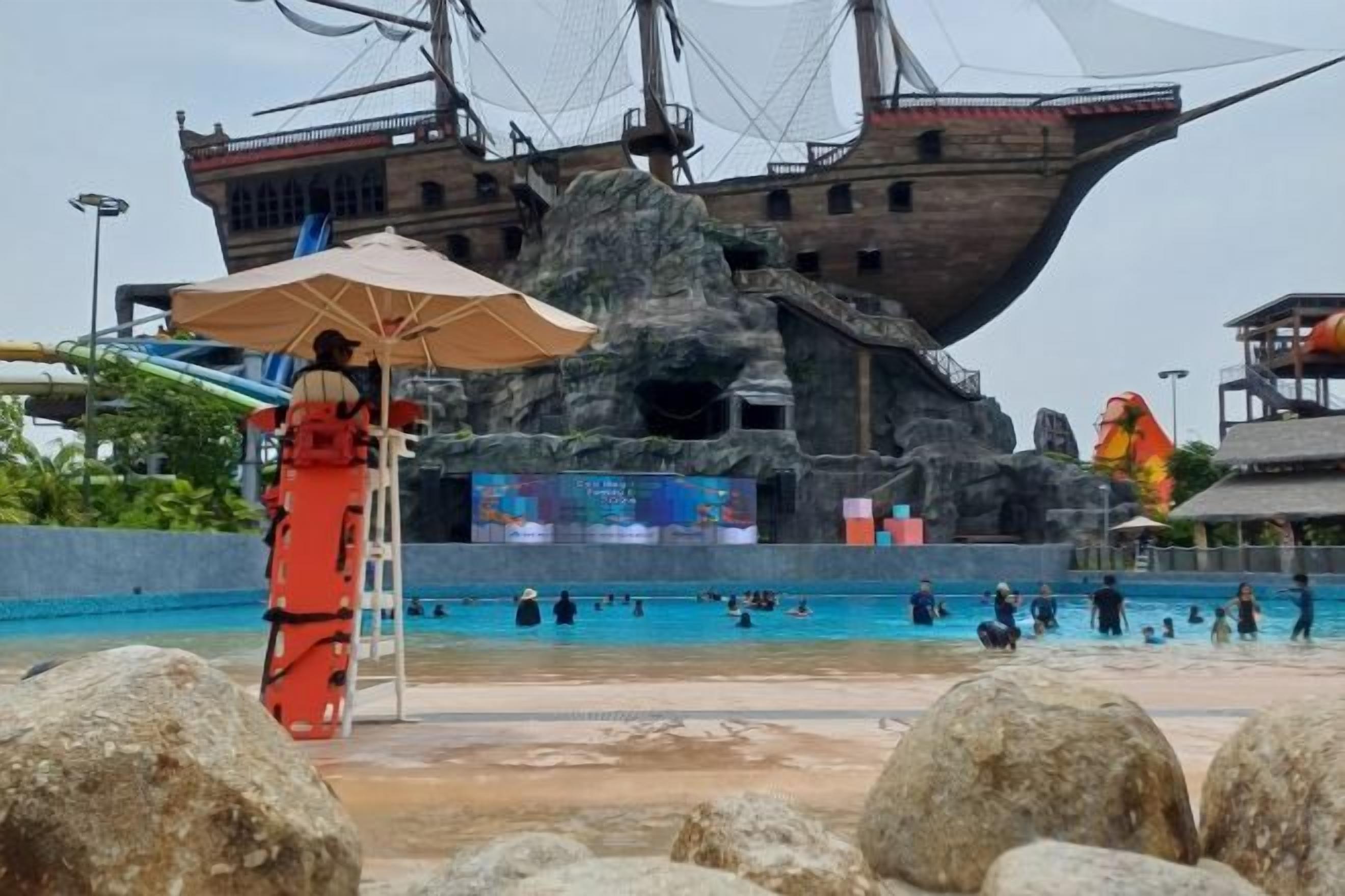 Una gran piscina exterior con un decorado con temática náutica, un puesto de socorrista y grandes rocas al frente.