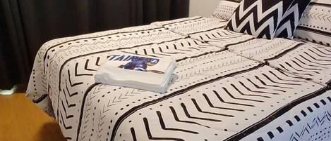 Una cama bien hecha con ropa de cama estampada y almohadas, una toalla doblada y un cabecero visible.